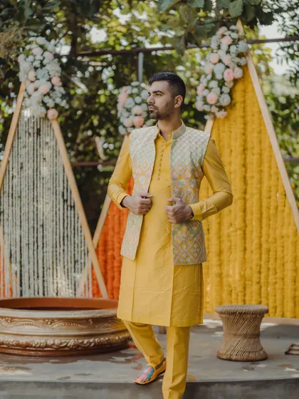 A groom poses against a modern Haldi backdrop made of triangular panels decorated with marigolds and white floral strings.
