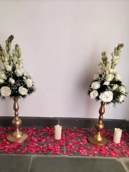 An elegant entrance detail featuring two golden pedestals with white floral arrangements. The floor is scattered with red rose petals, creating a romantic and welcoming pathway for guests.