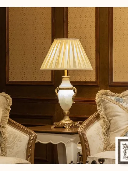 This classic table lamp combines a golden base with a white alabaster-style body, creating a look of pure elegance. It sits perfectly between two ornate, fabric-upholstered armchairs.