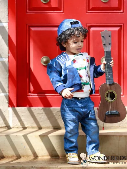 A little rockstar in the making! This toddler looks stylish in his denim outfit and cap, holding a guitar against a bright red door for a cool, urban-style kid's portrait.
