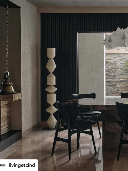 A detail from the dining area, showing the heavy stone table, modern black chairs, and a sculptural white floor lamp. The hanging temple bell adds a touch of Indian heritage.