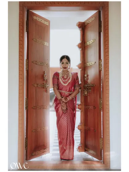A grand bridal entrance. This stunning portrait of the bride in a red silk saree, framed by magnificent traditional doors, creates a powerful and regal image.