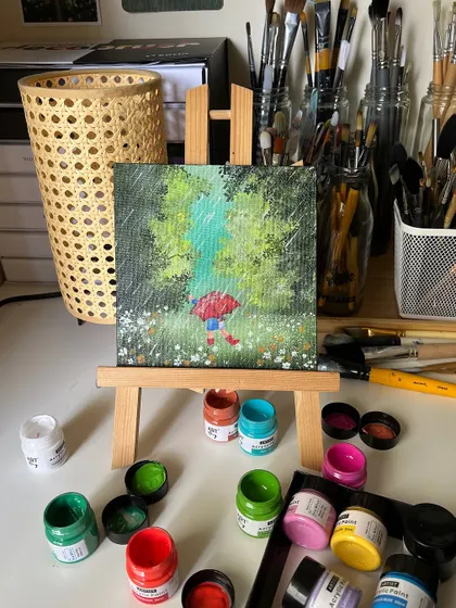 My rainy day painting on an easel, surrounded by the acrylic paints I used. The bright red umbrella adds a perfect pop of color to the green landscape.