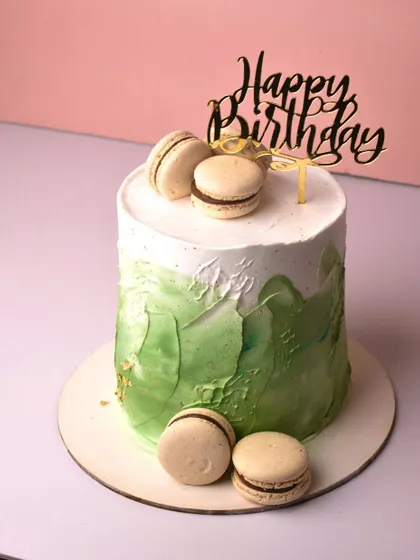 Name a better duo than cake and macarons. This tall cake features a modern green watercolor or spatula-painted effect, topped with a "Happy Birthday" sign and delicious chocolate-filled macarons.