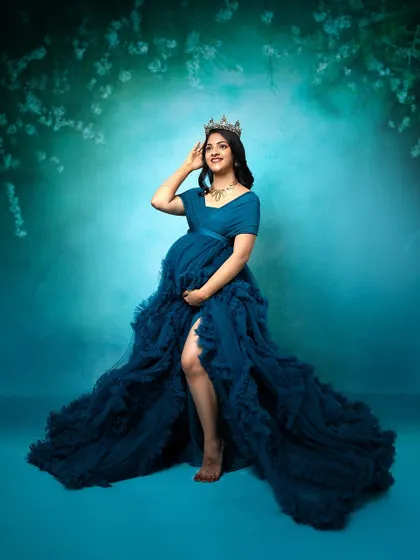 A glamorous maternity photoshoot. The makeup is elegant and bold, matching the dramatic teal gown and creating a stunning, queen-like portrait for the mother-to-be.