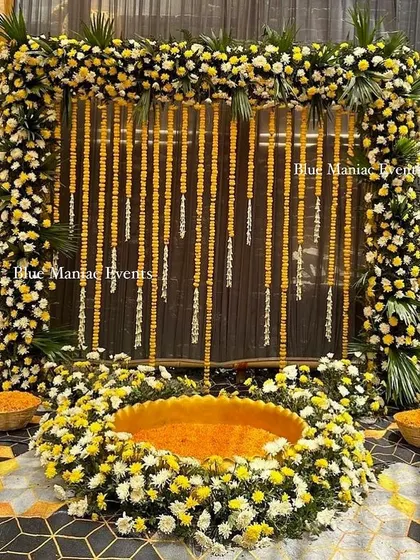 A luxurious Haldi setup using real flowers. The square frame is covered in marigolds and white chrysanthemums, with a flower-filled urli at the center for a truly opulent look.