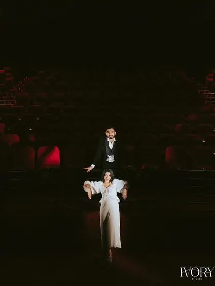 This wide shot uses dramatic lighting to create a powerful, minimalist composition in the theatre. The couple is the central focus in a vast, darkened space, symbolizing that they are the main characters in their own love story.