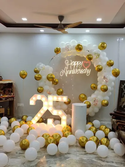 An elegant 25th-anniversary decoration in a living room. A white and gold balloon ring surrounds a neon sign, with a large light-up '25' and balloons scattered on the floor.