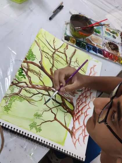 A close-up view shows the student adding fine details to the branches with a small brush, demonstrating precision in watercolor.