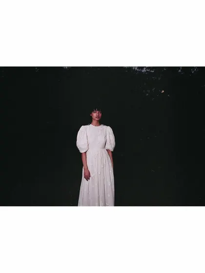 An atmospheric shot for the Sruti Dalmia campaign. The model is styled in a white dress with puff sleeves, positioned against a dark, natural background to create a moody and cinematic image that emphasizes solitude and form.