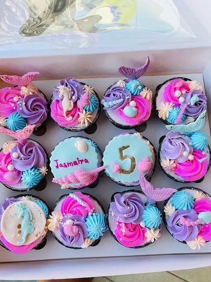 A closer look at the mermaid-themed cupcakes, featuring a mix of designs. Some have colorful frosting swirls with mermaid tail toppers, while others are personalized with the birthday girl's name and age.