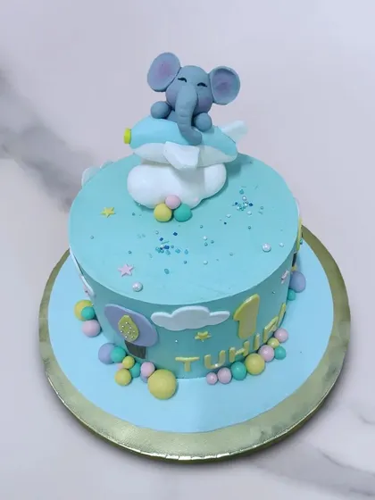 We fly high with this adorable elephant-themed first birthday cake. A cute baby elephant sits on a fluffy cloud, soaring through a pastel blue sky, making for a sweet and dreamy celebration of a special milestone.
