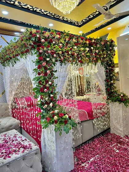 A luxurious wedding night setup with a bed framed by a dense arch of red and white flowers. A custom neon sign and hanging floral strings add to the glamour.