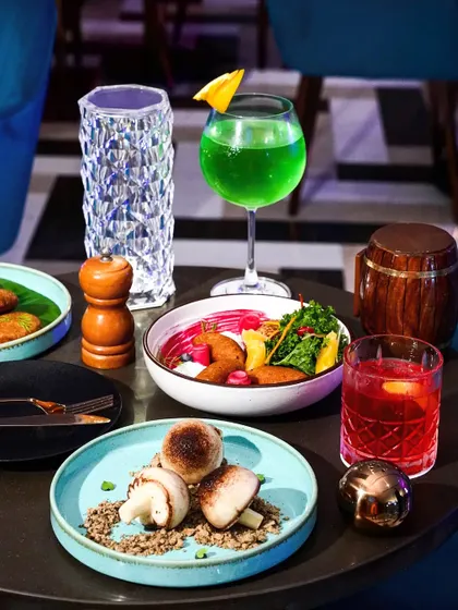 A table spread in a lounge, featuring appetizers like stuffed mushrooms and a bright green cocktail. The image captures the trendy and energetic vibe of the location.