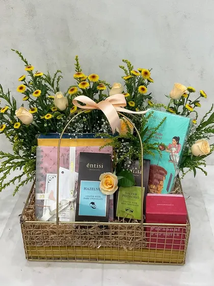 A classic Mother's Day gift basket. This golden wire basket is filled with books and chocolates, surrounded by a garden-style arrangement of yellow flowers and fresh greenery.
