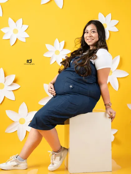 A bright and happy solo portrait. The cheerful yellow background and daisy props perfectly match the joyful smile of the mom-to-be in her casual denim outfit.
