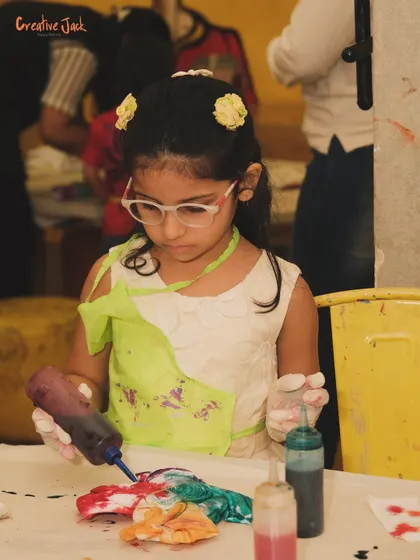 A young girl with glasses concentrates on her tie-dye creation, squeezing colorful dye onto a folded cloth.