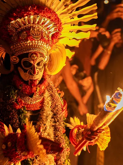 A close-up of the Karim Gullikan Theyyam, holding a torch that illuminates the intricate details of his mask and costume. The firelight reveals the textures and colors that bring the deity to life.