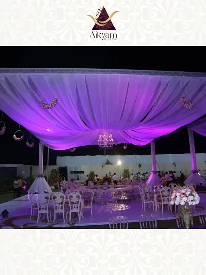 An expansive outdoor reception setup under a canopy of draped white fabric, illuminated by dramatic purple uplighting and a central crystal chandelier. The scene is set for a grand and elegant evening celebration.