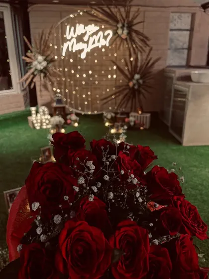 The final touch to a perfect question. A bouquet of deep red roses sits in the foreground of this boho-inspired "Will you Marry Me?" setup, complete with a jute backdrop and fairy lights.