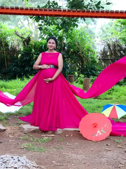 A full-length shot of a woman in a pink trail gown, posing in a park with colorful umbrellas in the background.
