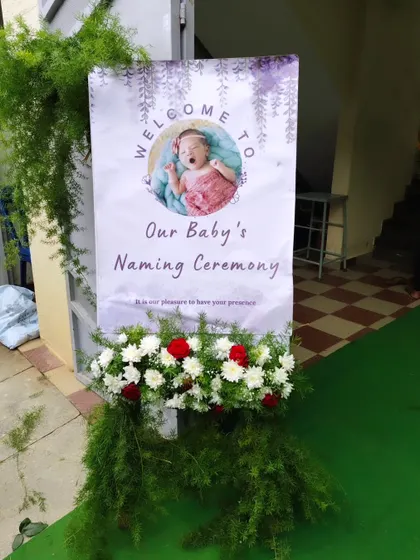 A personalized welcome board for a baby's naming ceremony. The stand is decorated with fresh red and white flowers.