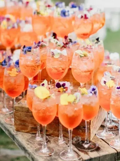 A large batch of Aperol Spritzes, garnished with edible flowers, ready for a wedding reception. This is a perfect welcome drink for guests, especially at a summer event.