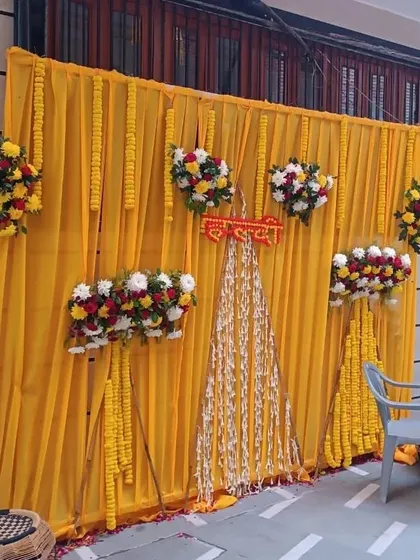 A simple Haldi backdrop for a home function, using a bright yellow drape accented with small floral bouquets and hanging marigold strings.