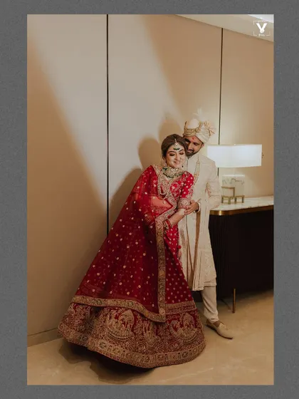 A full-length portrait of the couple in a hotel room, the bride's red lehenga adding a vibrant pop of color. The soft lighting and their relaxed pose create an intimate and elegant feel.