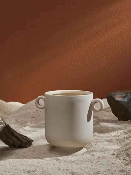 A white ceramic vase nestled in the sand. The contrast between the clean white ceramic and the rough textures of sand and wood creates a visually interesting composition.