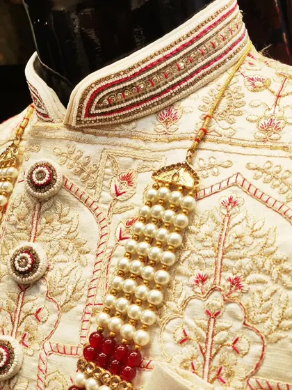 The difference is in the details. A close-up of a bespoke sherwani collar, showcasing intricate hand embroidery with red thread, pearls, and zardosi work on an ivory base, complete with custom-designed buttons.