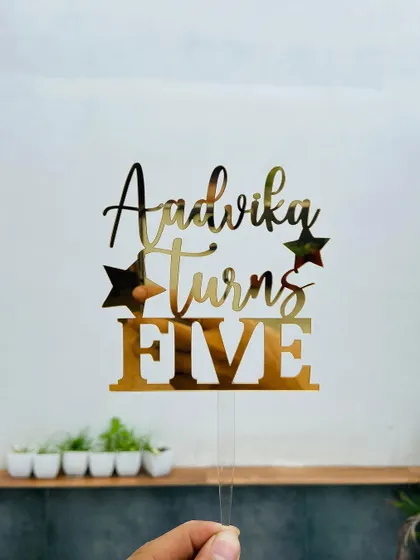 The "Aadvika Turns FIVE" topper held up against a bright background, showing off the clean cuts and reflective quality of the gold acrylic.