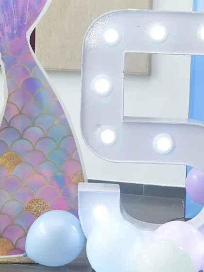A close-up of the details from a mermaid-themed party. This shot features a large, light-up marquee number '5' next to a custom-printed mermaid tail prop, surrounded by pastel balloons.