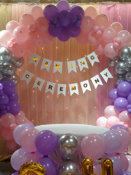 A close-up of the circular balloon backdrop for a naming ceremony, featuring a 'Naming Ceremony' banner inside a ring of pink, purple, and silver balloons.