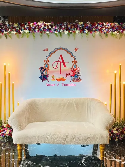 A simple and elegant stage for a Godhana and Chunadi ceremony. The white backdrop features a custom couple's monogram with traditional motifs, framed by floral arrangements and tall candles.