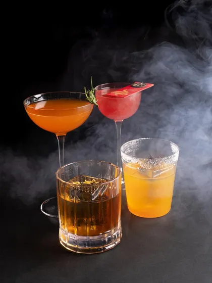A collection of four different cocktails, dramatically lit and surrounded by smoke for a moody and mysterious bar scene.
