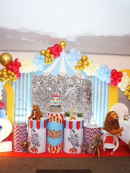 A classic carnival theme for Kashvik's first birthday. This setup includes a circus tent backdrop with a silver sequin curtain, themed pedestals, and red, blue, and gold balloon columns.