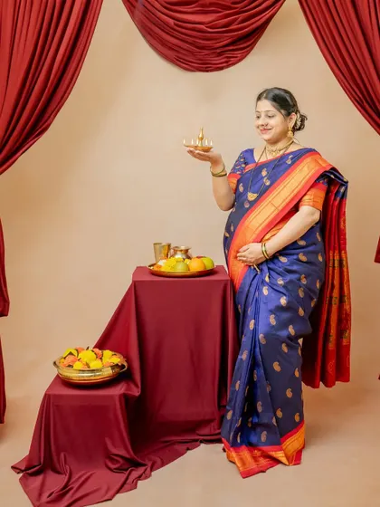 The mother-to-be is captured performing a traditional ritual with a diya. The rich red curtains and props create a warm, intimate, and sacred atmosphere for this 'Maatrutva' portrait.
