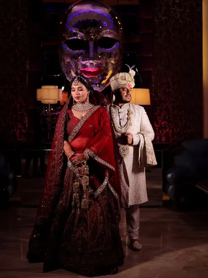 A dramatic, full-length portrait of the couple at their wedding venue. The unique mask sculpture in the background adds an artistic and modern touch.