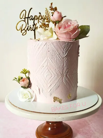 A tall, elegant pink cake with a modern, textured pattern. The cake is beautifully decorated with fresh flowers and a touch of gold leaf for a sophisticated finish.