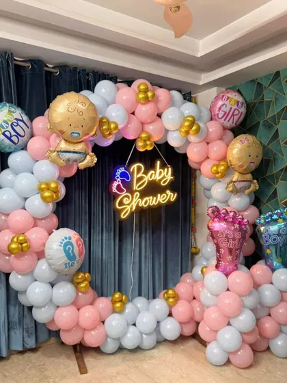 A popular 'He or She' themed baby shower decoration. This setup includes a circular balloon arch in pink and blue, with a central 'Baby Shower' neon sign and themed foil balloons.