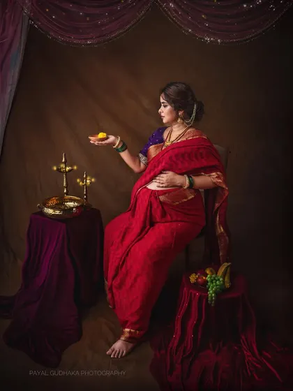 Inspired by the timeless paintings of Raja Ravi Varma, this studio portrait captures the divine grace of motherhood. Draped in a traditional red saree, this mother-to-be holds a 'laddoo', symbolizing the sweetness of new beginnings.