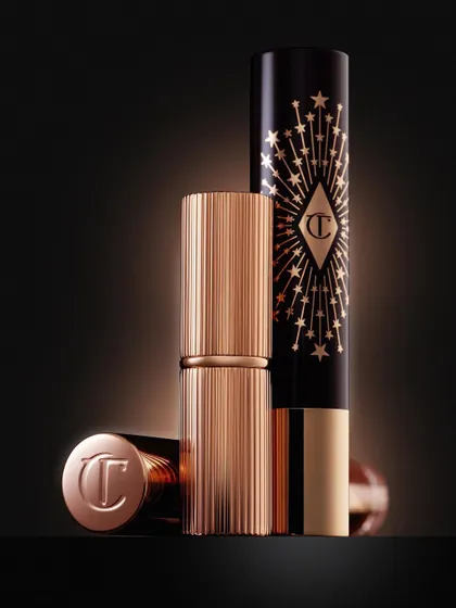 The texture and luxury of Charlotte Tilbury lipsticks are iconic. This shot focuses on the ribbed gold casing and the deep black packaging with its starburst design. The lighting is designed to make the gold gleam and feel incredibly premium.