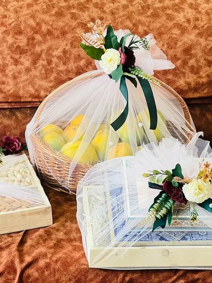 This trousseau set features a mix of fresh fruit baskets and gift boxes, all wrapped in white net with rich green and maroon ribbons. The presentation is traditional yet sophisticated.