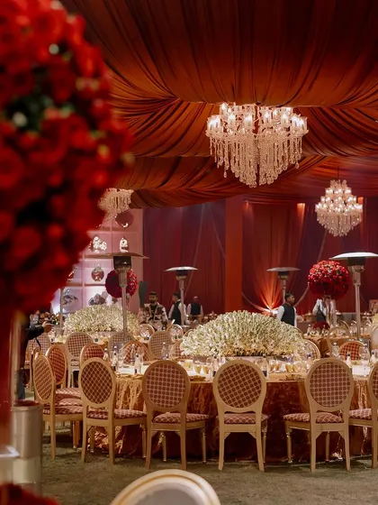 A wide view of the reception hall, blending traditional Indian opulence with modern luxury. A color palette of deep red and gold is accented with ivory florals, while dozens of chandeliers create a majestic atmosphere.