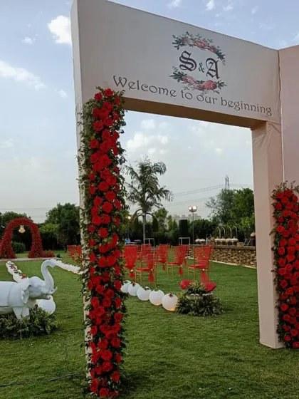 A grand and romantic wedding entrance. The aisle is lined with red chairs and leads to a stunning red floral arch, with a large "Welcome to our beginning" sign setting a beautiful tone for the ceremony.