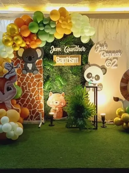 A cute woodland animals theme for a baptism and 2nd birthday celebration. The setup includes a faux grass floor, a balloon arch, and adorable cutouts of a deer, fox, lion, and panda.