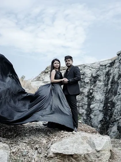 A wide shot capturing the scale of the landscape and the drama of the couple's pose, with her black gown flowing in the wind.