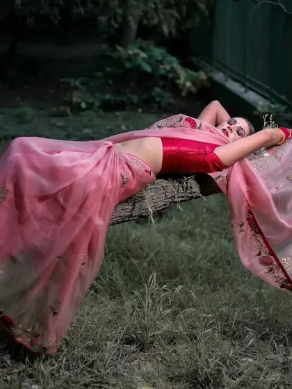 An artistic and dramatic pose, with the model lying on a fallen tree log in a lush green setting. The pink saree drapes beautifully, creating a stunning contrast with the natural surroundings.