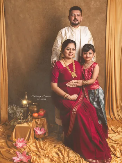 A regal family portrait celebrating a traditional Indian maternity journey. The mother, father, and daughter are dressed in elegant ethnic wear, surrounded by cultural props like brass vessels and lotuses, creating a timeless memory.